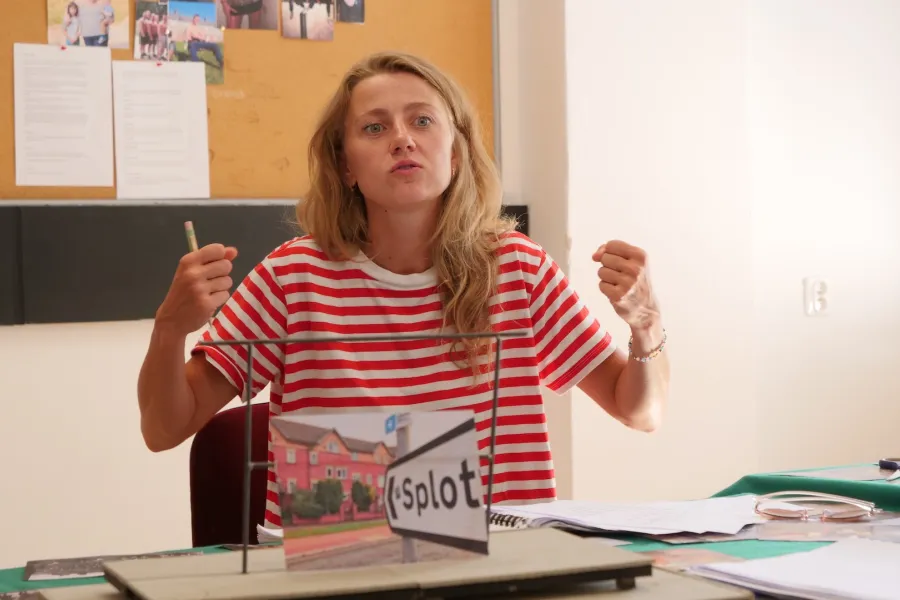 Zdjęcie przedstawia próbę stolikową do spektaklu „Ifigenia ze Splott”. Przy stole siedzi Agnieszka Skrzypczak z zgiętymi w łokciach i podniesionymi rękoma, których dłonie są zaciśnięte w pięści. Usta Agnieszki Skrzypczak są lekko ściągnięte i delikatnie otwarte. Przed aktorką na stole znajdują się fotografie i makieta osiedlowego trzepaka. Oprócz makiety na stole znajduje się zeszyt, okulary, scenariusz i nożyczki. 