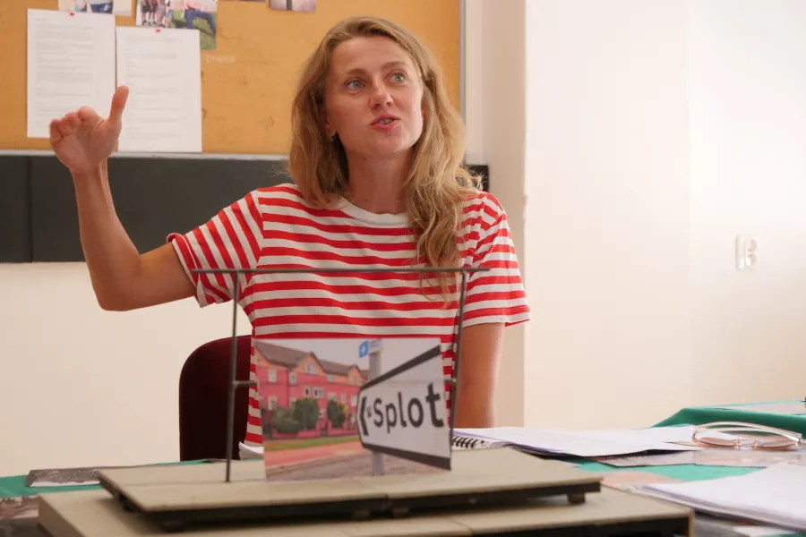 Zdjęcie przedstawia próbę stolikową do spektaklu „Ifigenia ze Splott”. Przy stole siedzi Agnieszka Skrzypczak z lewą ręką wyprostowaną a prawą podniesioną i zgiętą w łokciu. Dłoń tej ręki jest wyprostowana. Usta Agnieszki Skrzypczak są ściągnięte i delikatnie otwarte. Wzrok aktorki sięga ponad linię obiektywu. Przed aktorką na stole znajdują się fotografie i makieta osiedlowego trzepaka. Oprócz makiety na stole znajduje się zeszyt, okulary, scenariusz i nożyczki. 