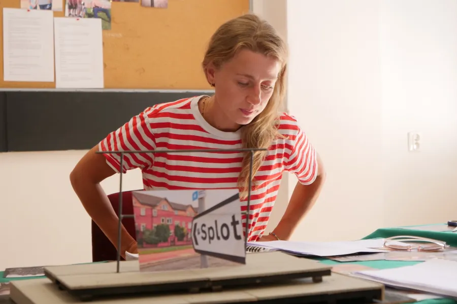 Zdjęcie przedstawia próbę stolikową do spektaklu „Ifigenia ze Splott”. Przy stole siedzi Agnieszka Skrzypczak z rękoma na biodrach. Wzrok aktorki utkwiony jest w scenariuszu a usta delikatnie ściągnięte do środka. Przed aktorką na stole znajdują się fotografie i makieta osiedlowego trzepaka. Oprócz makiety na stole znajduje się zeszyt, okulary, scenariusz i nożyczki. 