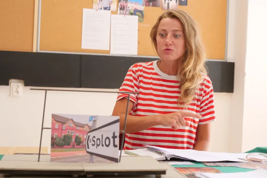 Zdjęcie przedstawia próbę stolikową do spektaklu „Ifigenia ze Splott”. Przy stole siedzi Agnieszka Skrzypczak. Wzrok aktorki utkwiony jest w makiecie trzepaka a jej dolna warga delikatnie wysunięta do przodu. Prawa ręka aktorki jest zgięta w łokciu i skierowana w bok, a wskazujący palec tej ręki jest wyprostowany. Przed aktorką na stole znajdują się fotografie i makieta osiedlowego trzepaka. 