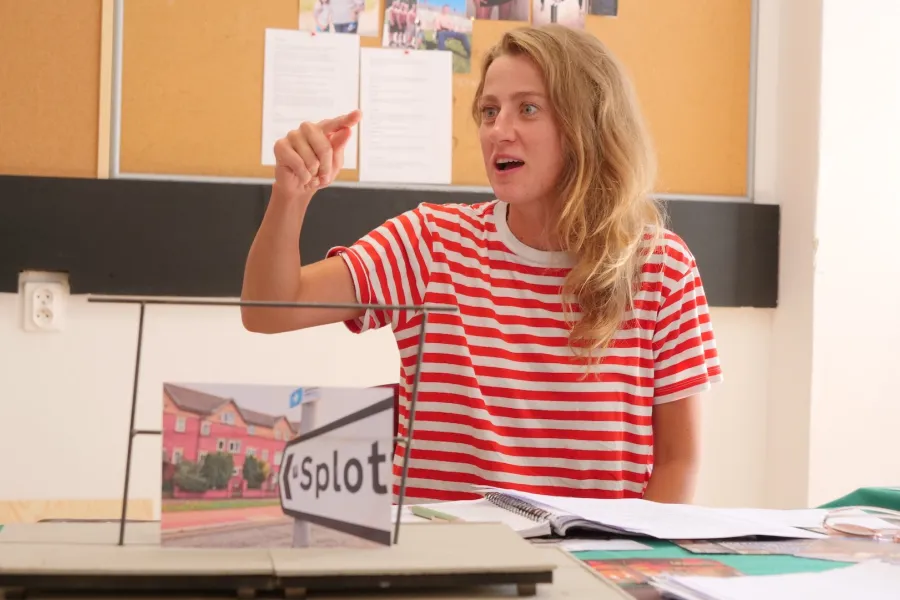 Zdjęcie przedstawia próbę stolikową do spektaklu „Ifigenia ze Splott”. Przy stole siedzi z lekko otwartymi ustami Agnieszka Skrzypczak. Wzrok aktorki utkwiony jest na wyprostowany palec wskazujący prawej, zgiętej w łokciu ręki. Przed aktorką na stole znajdują się fotografie i makieta osiedlowego trzepaka. 