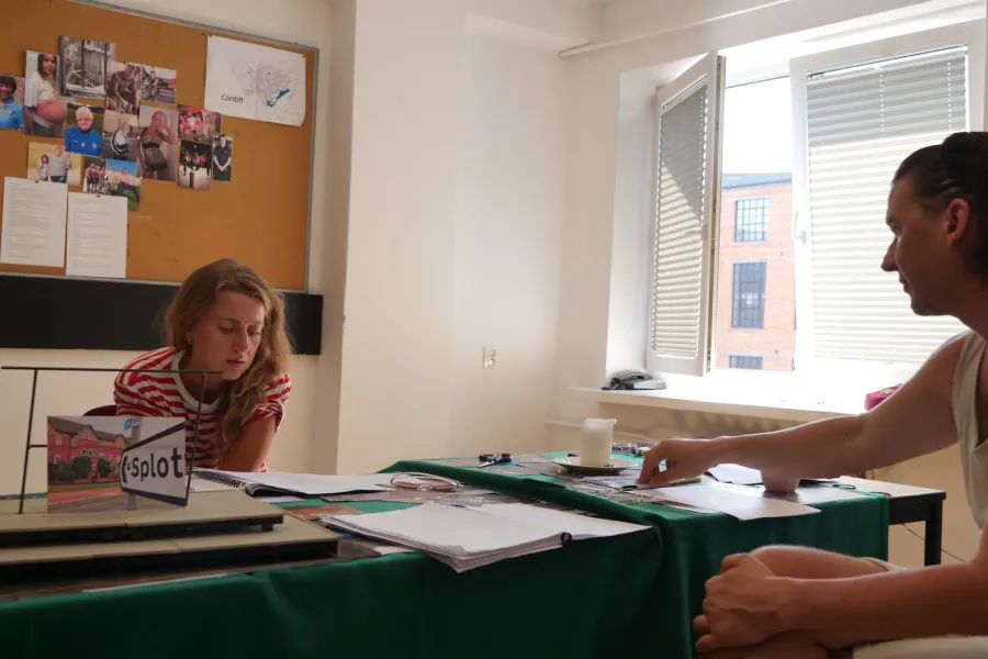 Zdjęcie przedstawia próbę stolikową do spektaklu „Ifigenia ze Splott”. Przy stole siedzi Agnieszka Skrzypczak i reżyser Filip Gieldon. Aktorka ze zgarbioną postawą patrzy w scenariusz przed sobą. Reżyser siedzi naprzeciwko aktorki, kierując swój wzrok na nią. Jego lewa ręka spoczywa na skrzyżowanych nogach a prawa na stole dociskając długopis. Na stole znajduje się makieta tapczanu ze zdjęciem angielskiej kamienicy i znakiem „Splott”, świeczka, okulary, nożyczki i scenariusze. Z tyłu wisi na ścianie tablica