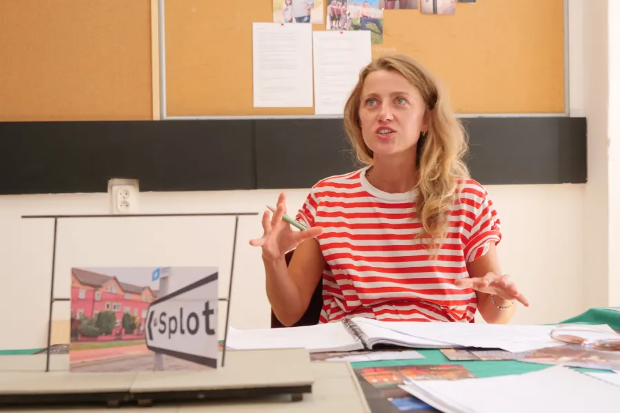 Zdjęcie przedstawia próbę stolikową do spektaklu „Ifigenia ze Splott”. Przy stole siedzi z lekko otwartymi ustami Agnieszka Skrzypczak. Wzrok aktorki utkwiony jest w przestrzeń powyżej linii obiektywu. Ręce Agnieszki Skrzypczak są zgięte w łokciach a palce dłoni mocno oddalone od siebie. Prawa ręka trzyma długopis. Przed aktorką na stole znajdują się fotografie, makieta osiedlowego trzepaka, okulary i scenariusz. Z tyłu widnieje tablica korkowa z tekstami i zdjęciami.