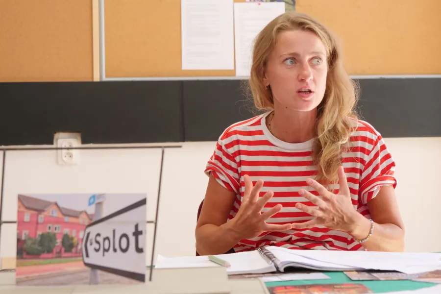 Zdjęcie przedstawia próbę stolikową do spektaklu „Ifigenia ze Splott”. Przy stole siedzi z lekko otwartymi ustami Agnieszka Skrzypczak. Wzrok aktorki utkwiony jest w przestrzeń po bocznej stronie obiektywu. Ręce Agnieszki Skrzypczak są zgięte w łokciach a palce dłoni mocno oddalone od siebie. Dłonie są zwrócone wewnętrzną częścią do aktorki. Przed aktorką na stole znajdują się fotografie, makieta osiedlowego trzepaka, okulary i scenariusz. Z tyłu widnieje tablica korkowa z tekstami i zdjęciami.