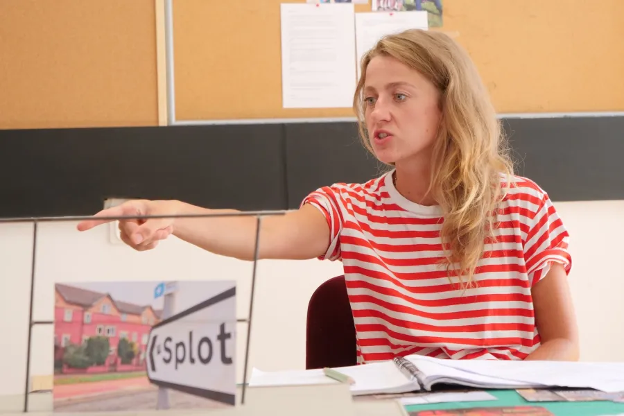 Zdjęcie przedstawia próbę stolikową do spektaklu „Ifigenia ze Splott”. Przy stole siedzi z lekko otwartymi ustami i zaciśniętymi zębami Agnieszka Skrzypczak. Wzrok aktorki utkwiony jest w przestrzeń poniżej linii obiektywu. Prawa ręka aktorki jest wyprostowana a palec wskazujący jest wyciągnięty do przodu. Przed aktorką na stole znajdują się fotografie, makieta osiedlowego trzepaka, okulary i scenariusz. Z tyłu widnieje tablica korkowa z tekstami i zdjęciami.