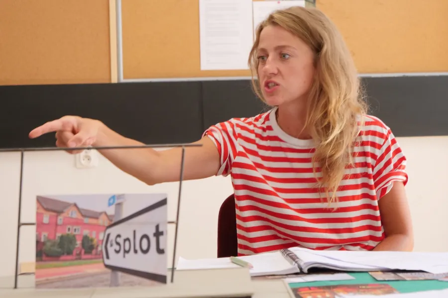 Zdjęcie przedstawia próbę stolikową do spektaklu „Ifigenia ze Splott”. Przy stole siedzi z lekko otwartymi ustami i zaciśniętymi zębami Agnieszka Skrzypczak. Wzrok aktorki utkwiony jest w przestrzeń poniżej linii obiektywu. Prawa ręka aktorki jest wyprostowana a palec wskazujący jest wyciągnięty do przodu. Przed aktorką na stole znajdują się fotografie, makieta osiedlowego trzepaka, okulary i scenariusz. Z tyłu widnieje tablica korkowa z tekstami i zdjęciami.