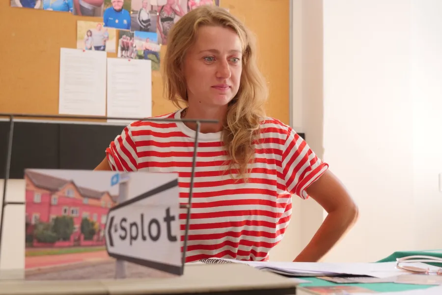 Zdjęcie przedstawia próbę stolikową do spektaklu „Ifigenia ze Splott”. Przy stole siedzi Agnieszka Skrzypczak z założonymi na biodra rękoma. Wzrok aktorki skierowany jest w przestrzeń, poniżej linii obiektywu. Przed Agnieszką Skrzypczak na stole znajdują się fotografie i makieta osiedlowego trzepaka z opartym na niej zdjęciem brytyjskiej kamienicy. Z tyłu na ścianie wisi tablica korkowa z różnymi zdjęciami i tekstami. 