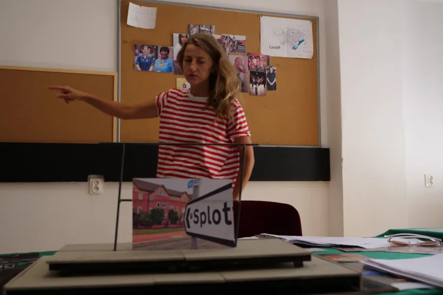 Zdjęcie przedstawia próbę stolikową do spektaklu „Ifigenia ze Splott”. Przed stołem z podniesioną prawą ręką i wyprostowanym palcem wskazującym stoi Agnieszka Skrzypczak. Natomiast lewa ręka aktorki spoczywa wzdłuż ciała. Wzrok aktorki jest spuszczony w dół, tak samo kącki ust. Na stole znajdują się fotografie i makieta osiedlowego trzepaka z opartym na niej zdjęciem brytyjskiej kamienicy. Z tyłu na ścianie wisi tablica korkowa z różnymi zdjęciami i tekstami. 