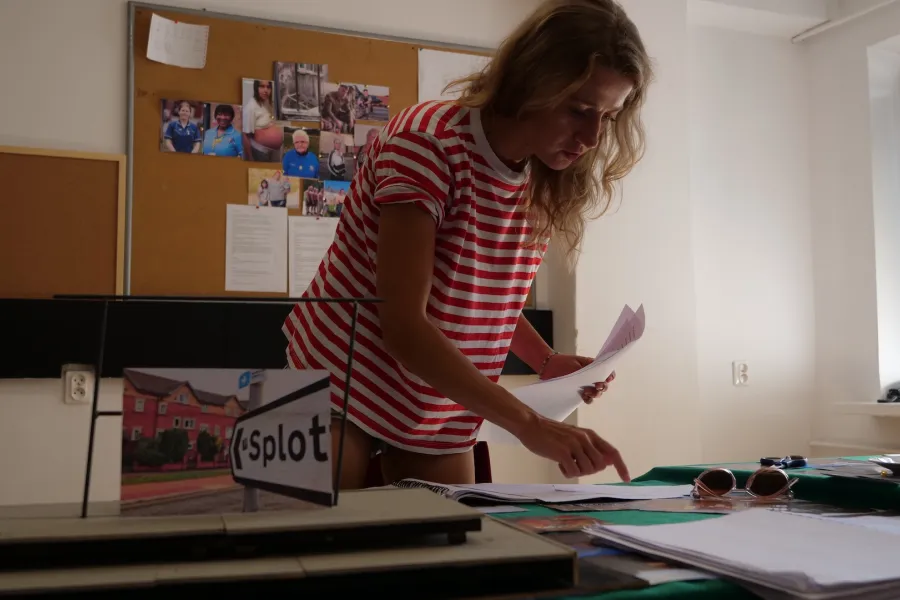 Zdjęcie przedstawia próbę stolikową do spektaklu „Ifigenia ze Splott”. Nad stołem pochyla się Agnieszka Skrzypczak. W lewej ręce trzyma kartki papieru, natomiast palcem wskazującym prawej ręki dotyka innych leżących kartek. Wzrok aktorki utkwiony jest na kartkach. Na stole znajdują się fotografie, okulary, scenariusz i makieta osiedlowego trzepaka z opartym na niej zdjęciem brytyjskiej kamienicy. Z tyłu na ścianie wisi tablica korkowa z różnymi zdjęciami i tekstami. 