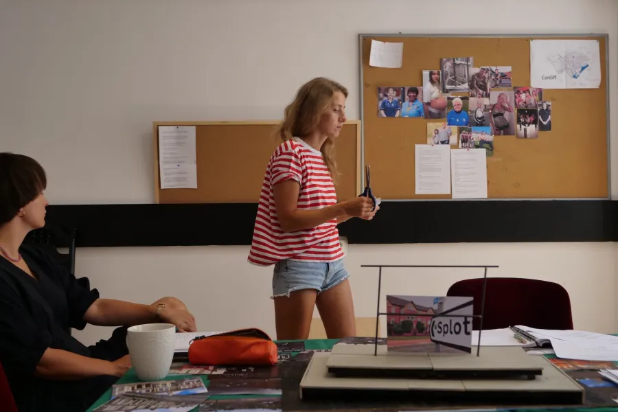 Zdjęcie przedstawia próbę stolikową do spektaklu „Ifigenia ze Splott”. Agnieszka Skrzypczak stoi bocznym profilem i patrzy na trzymane nożyczki i papier. Aktorka jest w centrum fotografii. Po lewej stronie aktorki wiszą na ścianie dwie tablice korkowe z tekstami i zdjęciami różnych osób. Przy stole, w lewym rogu zdjęcia, z ręką na kolanie siedzi Marta Baraszkiewicz. Stół jest na środku zdjęcia, po prawej stronie aktorki. Leży na nim kubek, piórnik, fotografie, makieta trzepaka z opartym na nim zdjęciem.