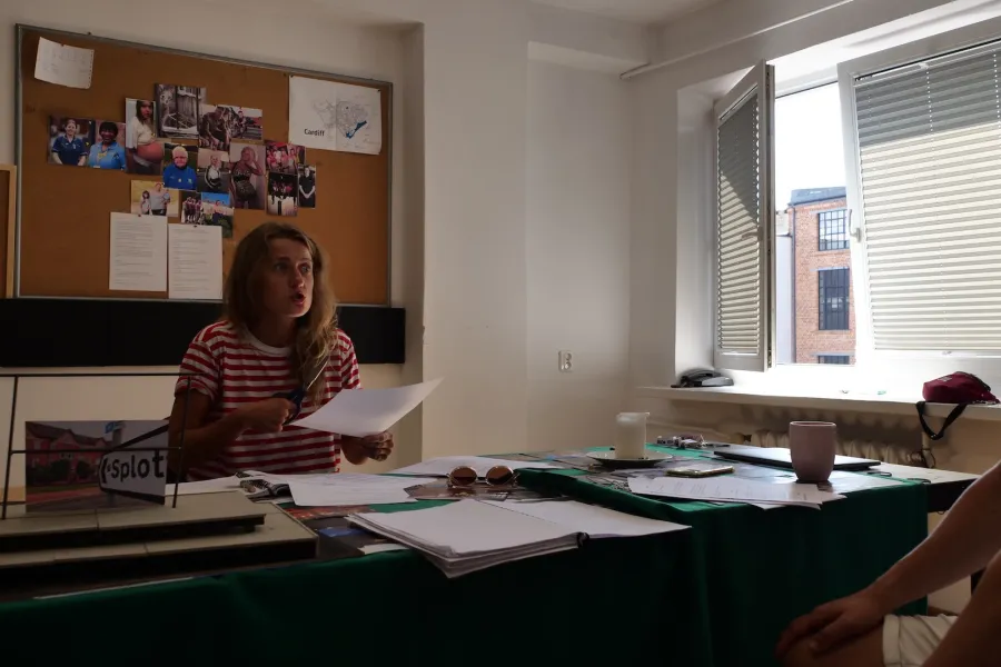 Zdjęcie przedstawia próbę stolikową do spektaklu „Ifigenia ze Splott”. Przy stole siedzi reżyser Filip Gieldon wraz z Agnieszką Skrzypczak. Reżyser siedzi z rękoma położonymi na swoich kolanach. Aktorka siedząc naprzeciwko reżysera trzyma w prawej ręce nożyczki a w lewej kartkę papieru. Wzrok Agnieszki Skrzypczak skierowany jest na reżyser. Jej oczy są mocno wytrzeszczone a usta ściągnięte i lekko otwarte.  Na stole znajdują się fotografie, okulary, świeca, kubek, scenariusz i makieta osiedlowego trzepaka.