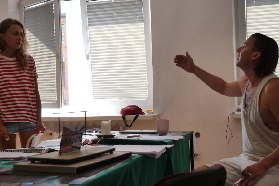 Zdjęcie przedstawia próbę stolikową do spektaklu „Ifigenia ze Splott”. Przy stole siedzi reżyser Filip Gieldon a naprzeciwko niego z rękami wzdłuż ciała stoi Agnieszka Skrzypczak. Reżyser podnosząc prawą rękę do góry utrzymuje kontakt wzrokowy z aktorką. Usta reżysera są otwarte a głowa pochylona lekko do tyłu. W swojej lewej ręce trzyma czarny długopis. Na stole znajdują się fotografie, okulary, świeca, kubek, scenariusz i makieta osiedlowego trzepaka z opartym na niej zdjęciem brytyjskiej kamienicy.