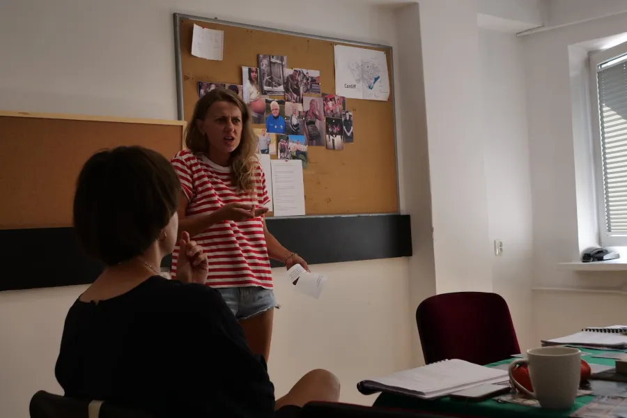 Zdjęcie przedstawia próbę stolikową do spektaklu „Ifigenia ze Splott”. Przy stole, tyłem do obiektywu siedzi inspicjentka Marta Baraszkiewicz a obok niej stoi Agnieszka Skrzypczak. Aktorka w lewej ręce trzyma tekst a prawą ma podniesioną na wysokość klatki. Jej brwi są zmarszczone, usta otwarte, ramiona podniesione do góry a wzrok skierowany przed siebie. Lewa ręka inspicjentki jest podniesiona na wysokość brody. Na stole znajduje się scenariusz, kubek, telefon i piórnik. Z tyłu wisi tablica krokowa. 