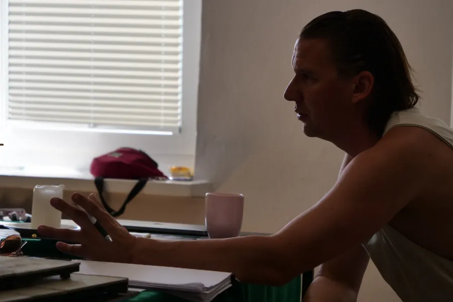 Zdjęcie przedstawia próbę stolikową do spektaklu „Ifigenia ze Splott”. Przy stole bocznym profilem siedzi reżyser Filip Gieldon. Lewa ręka trzymająca długopis leży na stole z lekko rozstawionymi od siebie palcami. Wzrok reżysera skierowany jest przed siebie a jego brwi są lekko zmarszczone. Na stole znajduje się makieta, świeca, kubek i scenariusz. 