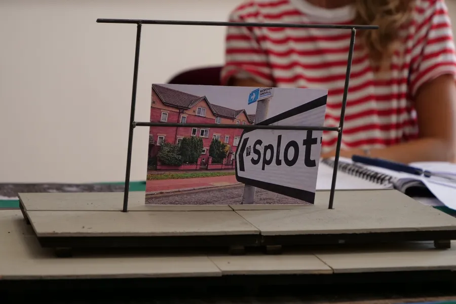 Zdjęcie przedstawia próbę stolikową do spektaklu „Ifigenia ze Splott”. Zdjęcie skoncentrowane na makiecie osiedlowego trzepaka, na którym opiera się fotografia brytyjskiej kamienicy, którą poprzedza znak z nazwą dzielnicy Splott. W tle został uchwycony tułów aktorki z jej koszulką w czerwono- białe paski. Na drugim planie można, również dostrzec scenariusz, nożyczki i fotografie.  