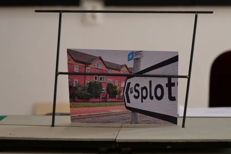 Zdjęcie przedstawia próbę stolikową do spektaklu „Ifigenia ze Splott”. Zdjęcie skoncentrowane na makiecie osiedlowego trzepaka, na którym opiera się fotografia brytyjskiej kamienicy, którą poprzedza znak z nazwą dzielnicy Splott. W tle został uchwycony tułów aktorki z jej koszulką w czerwono- białe paski. Na drugim planie można, również dostrzec scenariusz, nożyczki i fotografie.  