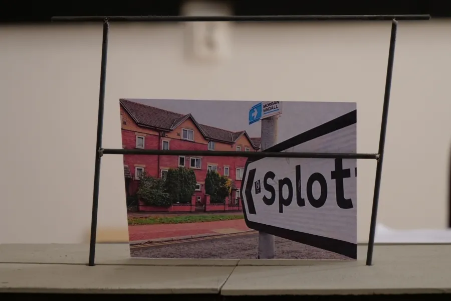 Zdjęcie przedstawia próbę stolikową do spektaklu „Ifigenia ze Splott”. Zdjęcie skoncentrowane na makiecie osiedlowego trzepaka, na którym opiera się fotografia brytyjskiej kamienicy, którą poprzedza znak z nazwą dzielnicy Splott.