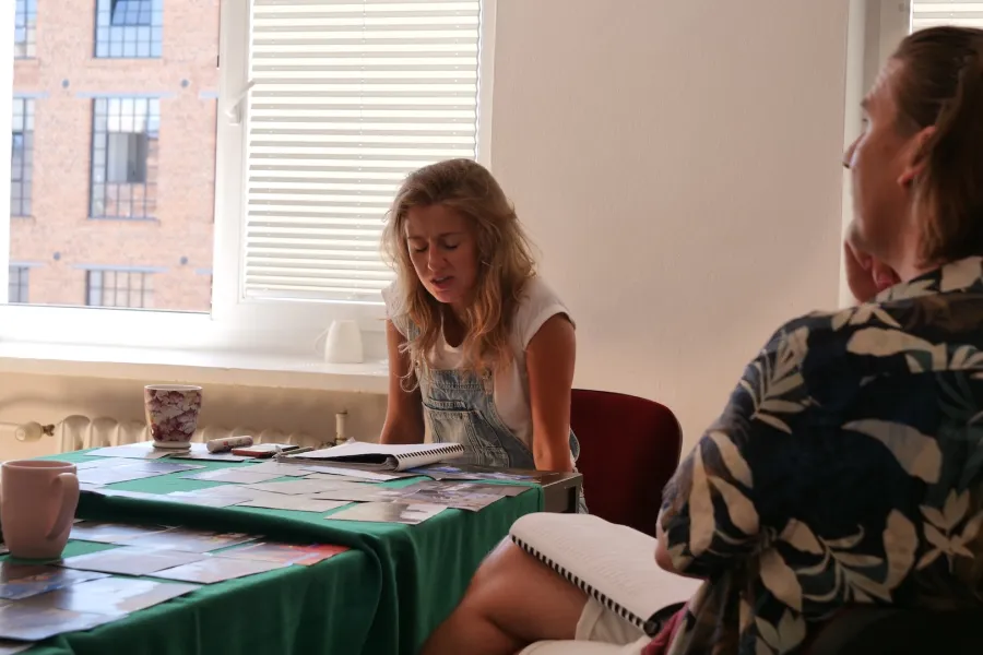 Zdjęcie przedstawia próbę stolikową do spektaklu „Ifigenia ze Splott”. Przy stole siedzi przodem Agnieszka Skrzypczak i tyłem reżyser Filip Gieldon. Na stole przed aktorką leży otwarty scenariusz i fotografie. Oczy aktorki są zamknięte a  ręce wyprostowane w łokciach, oparte o siedzenie. Reżyser trzyma na kolanach scenariusz a jego wzrok skierowany jest przed siebie. Na stole znajdują się także kubki, okulary i telefon. 