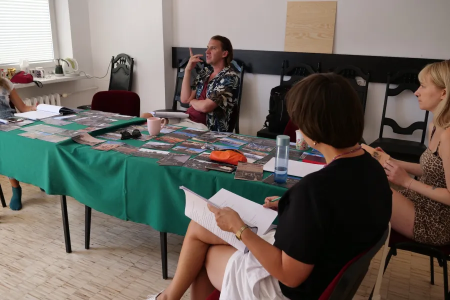 Zdjęcie przedstawia próbę stolikową do spektaklu „Ifigenia ze Splott”. ”. Przy stole siedzi od lewej strony trzymająca ręce na biodrach Agnieszka Skrzypczak, po środku z palcem wskazującym skierowanym ku górze reżyser Filip Gieldon, naprzeciwko aktorki scenografka i kostiumografka Adrianna Gołębiewska  i po prawej trzymająca scenariusz na nogach inspicjentka Marta Baraszkiewicz. Na stole leży dużo fotografii, scenariusz, piórnik, kubek i okulary. Przy stole leżą buty Agnieszki Skrzypczak i butelka soku.