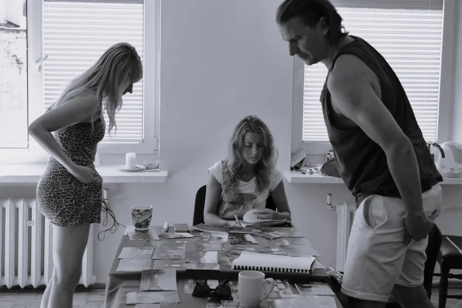 Zdjęcie przedstawia próbę stolikową do spektaklu „Ifigenia ze Splott”. Zdjęcie jest czarno-białe.  Przy stole siedzi Agnieszka Skrzypczak, która przegląda scenariusz. Nad tym samym stołem pochyla się reżyser Filip Gieldon wraz z scenografką Adrianną Gołębiewską. Spoglądają oni na rozłożone na stole zdjęcia. Oboje są skierowani bokiem do obiektywu, po środku nich przodem siedzi Agnieszka Skrzypczak. Na stole oprócz zdjęć znajdują się kubki, okulary i telefon. 