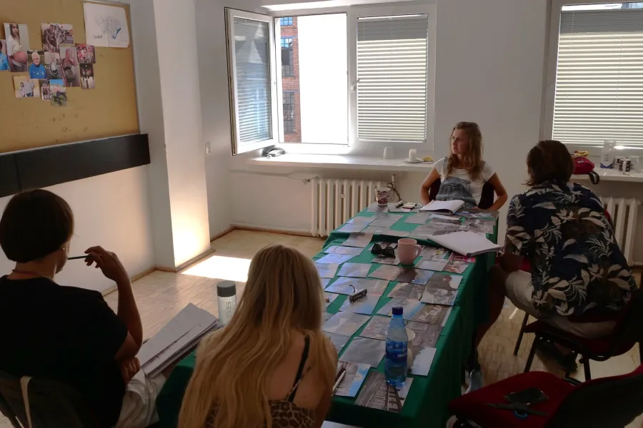 Zdjęcie przedstawia próbę stolikową do spektaklu „Ifigenia ze Splott”. Fotografia obejmuje dużą część pomieszczenia w tym cały stół, przy którym siedzi reżyser Filip Gieldon, inspicjentka Marta Baraszkiewicz, Agnieszka Skrzypczak i scenografka Adrianna Gołębiewska. Aktorka, która spogląda na tablicę korkową jako jedyna jest przodem do obiektywu, natomiast pozostałe osoby są zwrócone tyłem. Na stole leży dużo fotografii, kubki, butelki i scenariusze