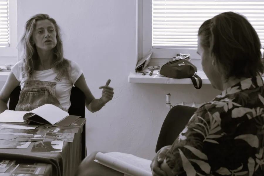 Zdjęcie przedstawia próbę stolikową do spektaklu „Ifigenia ze Splott”. Zdjęcie jest czarno- białe. Przy stole siedzi przodem Agnieszka Skrzypczak i tyłem reżyser Filip Gieldon. Na stole przed aktorką leży otwarty scenariusz i fotografie. Usta aktorki są lekko otwarte i głowa lekko pochylona do tyłu. Lewa ręka jest lekko wyciągnięta do boku a palec wskazujący i kciuk tej ręki wyprostowany. Kontakt wzrokowy skierowany jest na reżysera, który na swoich kolanach trzyma scenariusz. 