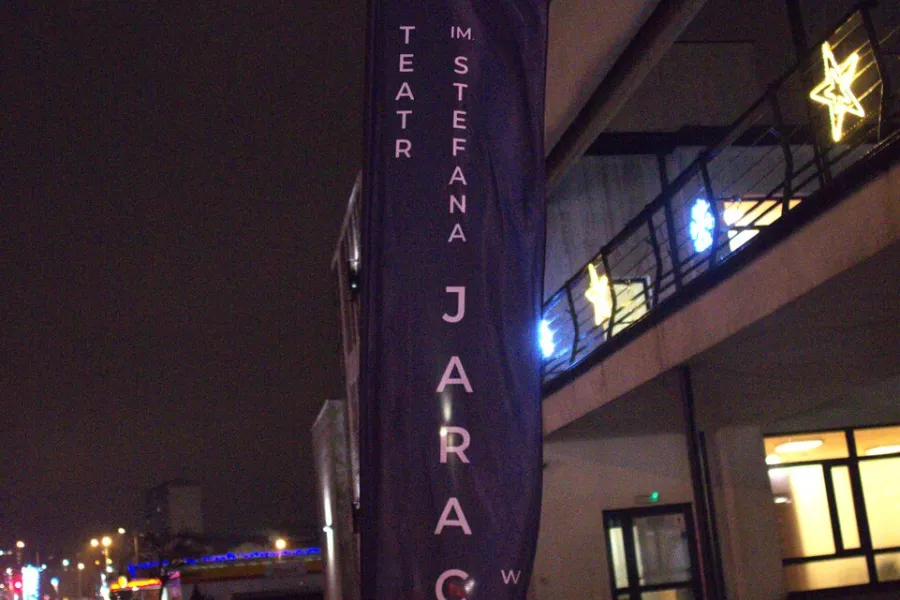 zdjęcie przedstawia flagi Teatru Jaracza w Łodzi z napisem z nazwa instytucji. Flagi są duże, umieszczone na stelarzach przed wejściem do budynku MDK w Radomsku. 