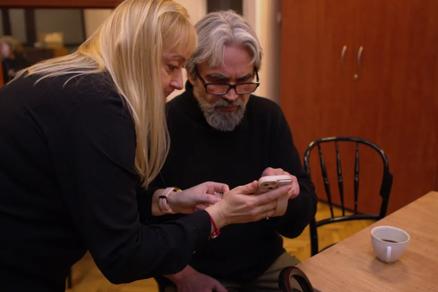 Zdjęcie przedstawia inspicjentkę, która pokazuje aktorowi Dariuszowi Bereskiemu coś na telefonie komórkowym. Zdjęcie zostało wykonane w garderobie teatralnej. Aktor siedzi przy stole, inspicjentka pochyla się nad nim. 