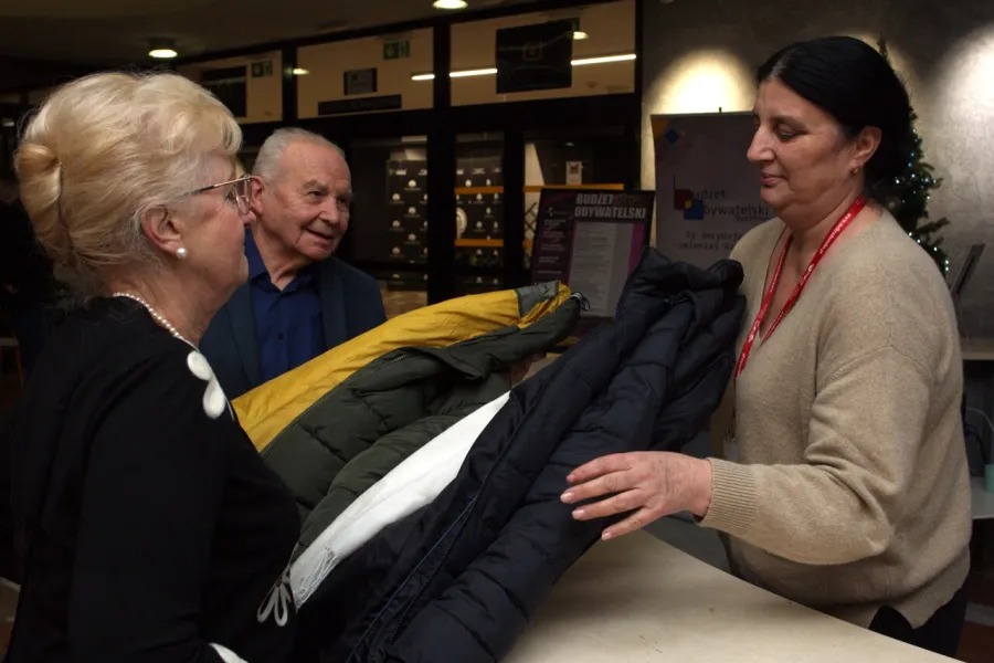 Zdjęcie przedstawia dwójkę widzów oddających do szatni swoje kurtki, które odbiera szatniarka. 