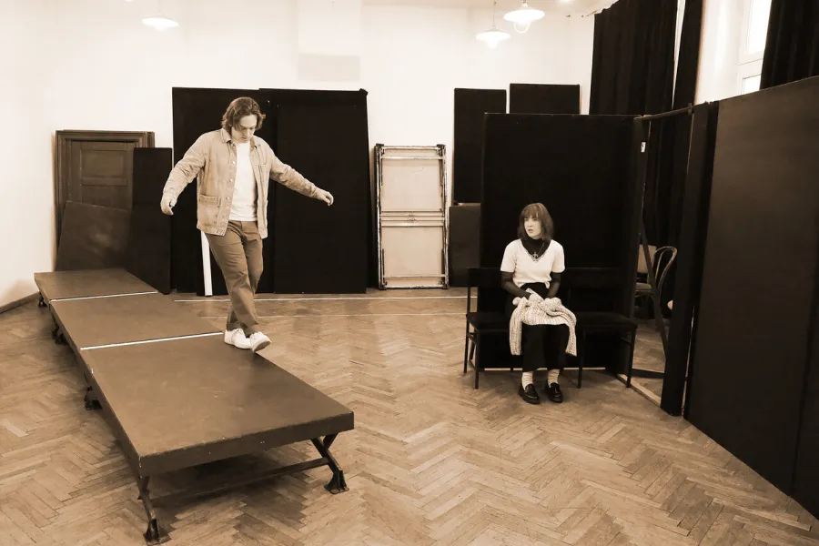 Zdjęcie przedstawia dwójkę aktorów. Aktor stoi na krawędzi podestu, balansując. W tle siedzi aktorka Szantal Strzelińska. 