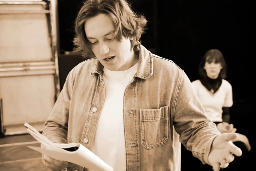 zdjęcie przedstawia aktora Konrada Krupińskiego, który trzyma w dłoniach scenariusz sztuki. Za nim w tle stoi aktorka Szantal Strzelińska. 