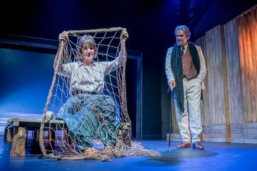 na zdjęciu aktorka Szantal Strzelińska siedzi na drewnianym pomoście, w rękach trzyma rybacką sieć, przygotowuje się do jej szycia. Po jej lewej stronie stoi aktor Dariusz Bereski i przygląda się jej