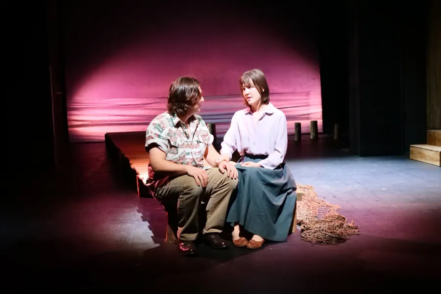 Zdjęcie z próby medialnej spektaklu „Zamiana". Zdjęcie przedstawia scenę ze spektaklu „Zamiana". Dwoje aktorów Konrad Krupiński i Szantal Strzelińska, siedzą na drewnianym podeście. W tle jest różowo-czerwono-białe światło imitujące zachód słońca nad morzem. 