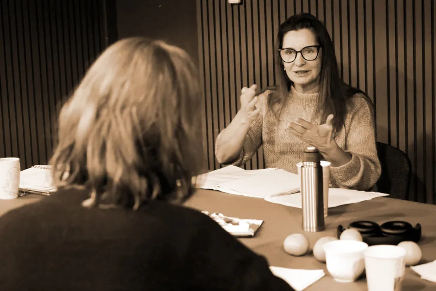 Zdjęcie z prób do spektaklu „Zamiana". Aktorka siedzi za stołem, na którym leżą scenariusze sztuki. Tyłem na drugim krześle siedzi druga aktorka.