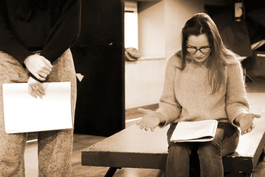 Zdjęcie z próby do spektaklu „Zamiana". Na zdjęciu widać jedną z aktorek od pasa w dół, w rękach trzyma scenariusz. Druga z nich siedzi na podeście, na kolanach ma scenariusz sztuki.
