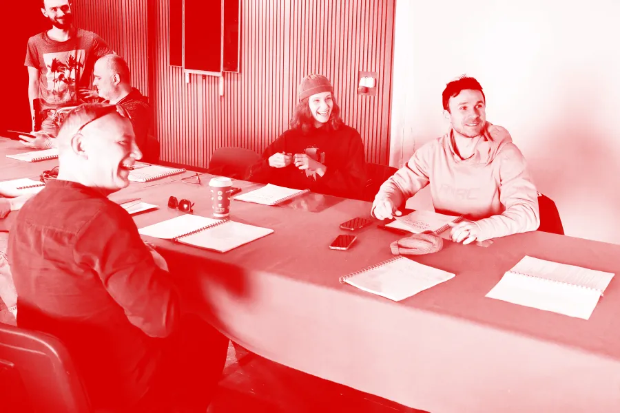 zdjęcie przedstawia pierwszą próbę spektaklu "Morderstwo w hotelu". Przy stole siedzą aktorzy.Tyłem do obiektywu siedzi śmiejący się aktor Mariusz Witkowski. Po prawej stronie stołu, przy stole stoi pracownik teatru Marcin Majczak. Na krzesłach obok siedzą scenograf Grzegorz Policiński,aktorzy Natalia Klepacka i Krzysztof Wach. Na stole znajdują się egzeplarze tekstu. Na zdjęcie nałożony został czerwony filtr. 