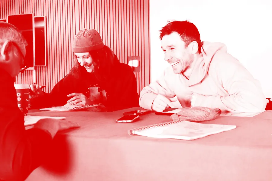 Zdjęcie przedstawia pierwszą próbę spektaklu „Morderstwo w hotelu". Po lewej stronie stołu siedzi aktor Mariusz Witkowski. Na przeciwko niego, po drugiej stronie stołu siedzą aktorzy Natalia Klepacka i Krzysztof Wach. Aktorka ma na głowie czapkę. Wszyscy aktorzy się śmieją. Na zdjęcie został nałożony czerwony filtr.