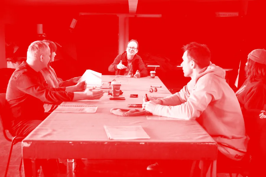 zdjęcie przedstawia pierwszą próbę spektaklu "Morderstwo w hotelu". Przy stole siedzą aktorzy, inspicjentka i reżyser. Po lewej stronie siedzą aktor Mariusz Witkowski i inspicjentka Marta Baraszkiewicz, po środku na szczycie stołu siedzi reżyser Jacek Bończyk, który coś tłumaczy. Po prawej stronie siedzą aktorzy Natalia Klepacka i Krzysztof Wach. Na stole znajdują się egzeplarze tekstu. Na zdjęcie nałożony został czerwony filtr.