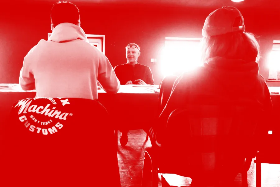 Zdjęcie przedstawia pierwszą próbę spektaklu „Morderstwo w hotelu". Tyłem do obiektywu siedzą przy stole aktorzy Krzysztof Wach i Natalia Klepacka (ona w czapce na głowie). Przodem do obiektywu, po drugiej stronie stołu siedzi aktor Mariusz Witkowski. Za nim znajdują się dwa okna, przez które przebija mocne światło słoneczne. Na zdjęcie został nałożony czerwony filtr. 