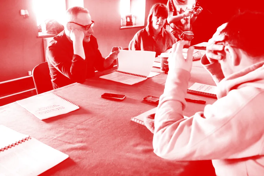 zdjęcie przedstawia pierwszą próbę spektaklu „Morderstwo w hotelu". Po lewej stronie stołu siedzi aktor Mariusz Witkowski czytający egzemplarz sztuki i przerzuca jego kartkę. Obok siedzi inspicjentka Marta Baraszkiewicz. W tle widać pracownika teatru Marcina Majczaka. Po prawej stronie blisko obiektywu siedzi aktor Krzystzof Wach zasłaniający prawą ręką swoją głowę. Lewą rekę ma uniesioną. Na zdjęcie został nałożony czerwony filtr. 