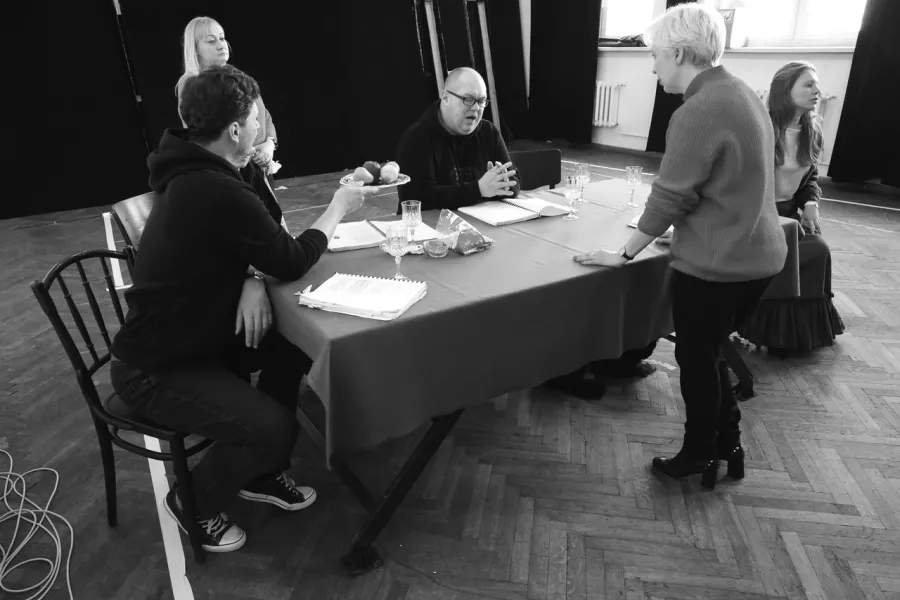 zdjęcie przedstawia próbę spektaklu „Świętoszek". Zostało zrobione na dużej sali prób w Teatrze Jaracza w Łodzi. Aktorzy Marcin Włodarski i Paweł Tucholski siedzą przy stole. Marcin Włodarski podaje Pawłowi Tucholskiemu tacę z owocami. Na stole są puste kieliszki do wina, które są rekwizytami w spektaklu oraz egzemplarze scenariusza. W tle widać stojącą inspicjentkę Katarzynę Brudek. Na krześle z boku przy stole siedzi aktorka Matylda Paszczenko. Nad stołem stoi reżyserka Magdalena Piekorz