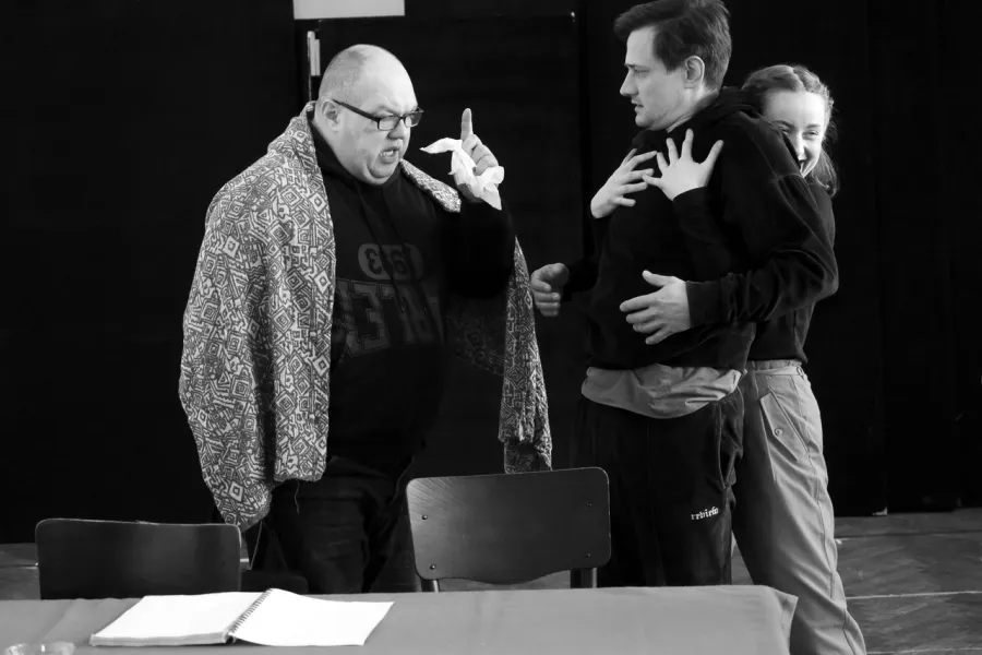 zdjęcie przedstawia próbę spektaklu „Świętoszek". Zostało zrobione na dużej sali prób w Teatrze Jaracza w Łodzi. Bokie do aparatu, za stołem stoi aktor Paweł Tucholski z uniesioną dłonią, w której trzyma chusteczkę. Na przeciwko niego stoi aktor Marek Nędza, za nim stoi aktorka Iwona Dróżdż, która go przytula od tyłu, trzymając dłonie na jego klatce piersiowej. 