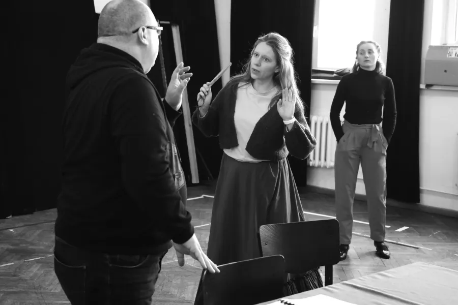 zdjęcie przedstawia próbę spektaklu „Świętoszek". Zostało zrobione na dużej sali prób w Teatrze Jaracza w Łodzi. Tyłem do aparatu stoi aktor Paweł Tucholski. Na przeciwko niego stoi aktorka Matylda Paszczenko, w ręku trzyma zwinięty wachlarz. W tle stoi aktorka Iwona Dróżdż.