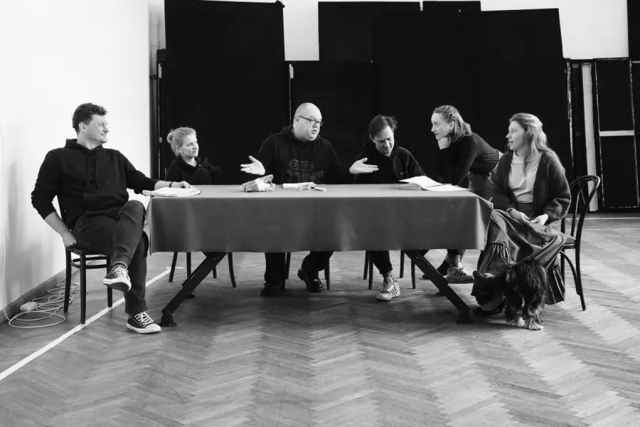 zdjęcie przedstawia próbę spektaklu „Świętoszek". Zostało zrobione na dużej sali prób w Teatrze Jaracza w Łodzi. Szóstka aktorów (od lewej: Marcin Włodarski, Sara Lityńska, Paweł Tucholski, Marek Nędza, Iwona Dróżdż, Matylda Paszczenko) siedzi przy stole przykrytym suknem. Wszyscy aktorzy patrzą na Pawła Tucholskiego, ten ma rozłożone ręce na boki, zgięte w łokciu. Iwona Dróżdż jako jedyna nie siedzi przy stole na krześle, a opieka się o niego łokciami. Zdjęcie jest czarno-białe.