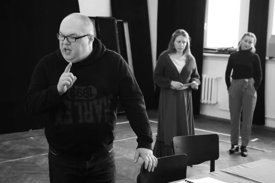 zdjęcie przedstawia próbę spektaklu „Świętoszek". Zostało zrobione na dużej sali prób w Teatrze Jaracza w Łodzi. Przodem do aparatu stoi Paweł Tucholski. Prawą dłoń ma uniesioną pokazując palec wskazujący. W tle stoją aktorki Matylda Paszczenko i Iwona Dróżdż