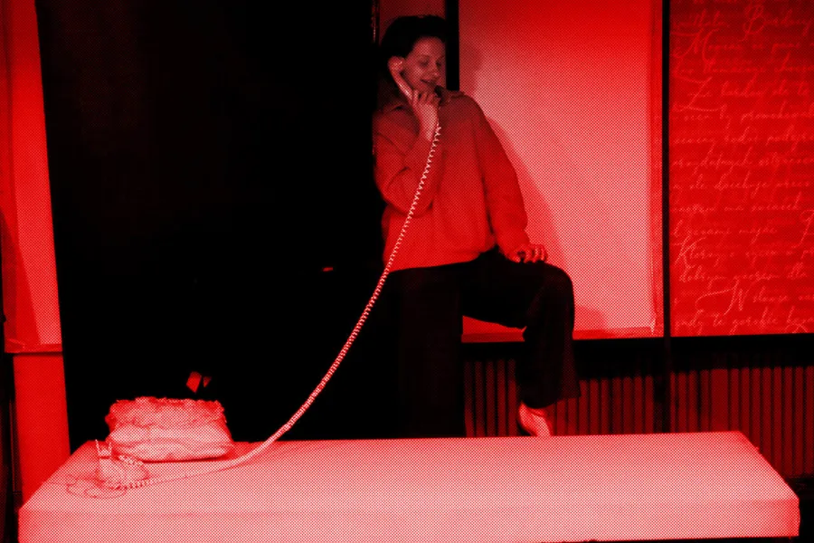 próba do spektaklu „Morderstwo w hotelu", zdjęcie zostało zrobione w sali prób, na zdjęciu: Natalia Klepacka, która stoi za łóżkiem i rozmawia przez telefon stacjonarny, którego słuchawka jest na sprężynowym, długim kablu, fot. Marcin Nowak. Na zdjęcie nałożony jest czerwony filtr. 