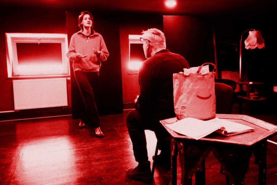 próba do spektaklu „Morderstwo w hotelu", zdjęcie zostało zrobione w sali prób, na zdjęciu: Natalia Klepacka idąca w kierunku siedzącego na krześle Mariusza Witkowskiego. Na niskim stoliku stoi papierowa torba z jedzeniem, obok leży scenariusz przedstawienia, fot. Marcin Nowak. Na zdjęcie nałożony jest czerwony filtr.