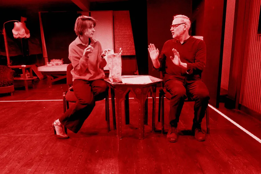 próba do spektaklu „Morderstwo w hotelu", zdjęcie zostało zrobione w sali prób, na zdjęciu: Natalia Klepacka, Mariusz Witkowski. Aktorzy siedzą na krzesłach przy niskim stoliku. Natalia Klepacka wyjmuje jedzenie z papierowej torby, fot. Marcin Nowak. Na zdjęcie nałożony jest czerwony filtr.
