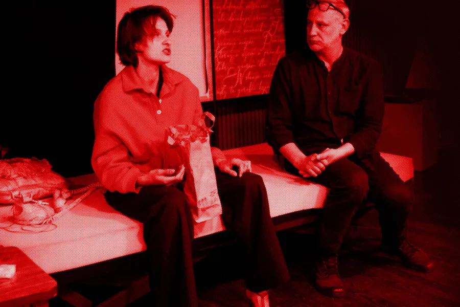 próba do spektaklu „Morderstwo w hotelu", zdjęcie zostało wykonane w sali prób, na zdjęciu: Natalia Klepacka, Mariusz Witkowski. Aktorzy siedzą obok siebie na łóżku. Aktorka ma między nogami papierową torbę z jedzeniem. Twarz ma zwróconą w przód. Aktor patrzy na nią, fot. Marcin Nowak. Na zdjęcie nałożony jest czerwony filtr. 