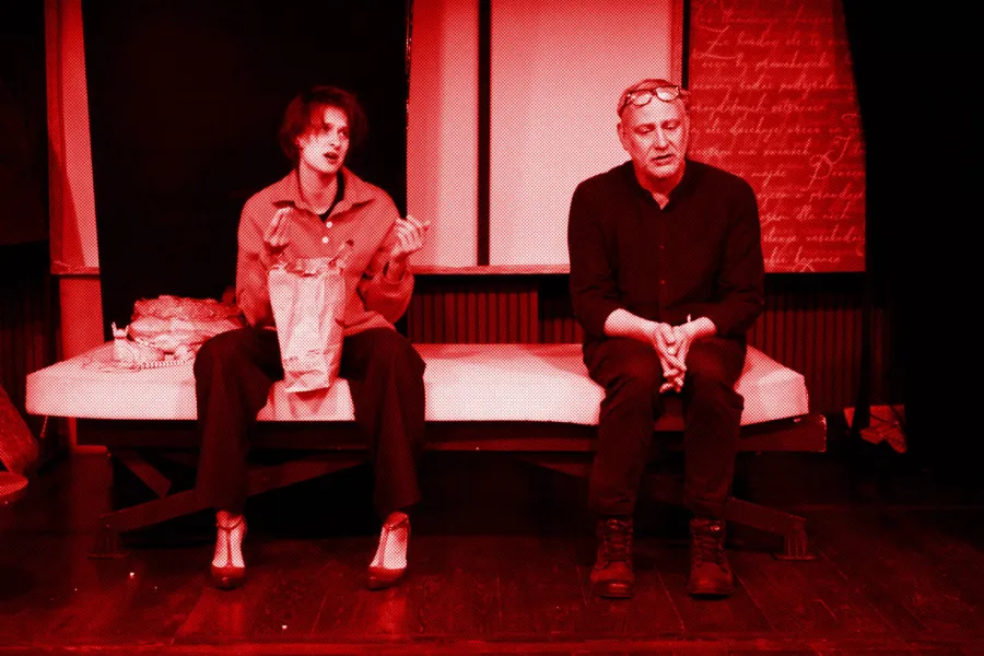 próba do spektaklu „Morderstwo w hotelu", zdjęcie zostało wykonane w sali prób, na zdjęciu: Natalia Klepacka, Mariusz Witkowski. Aktorzy siedzą obok siebie na łóżku. Aktorka ma między nogami papierową torbę z jedzeniem, siedzą twarzami zwróconymi do przodu, fot. Marcin Nowak. Na zdjęcie nałożony jest czerwony filtr. 